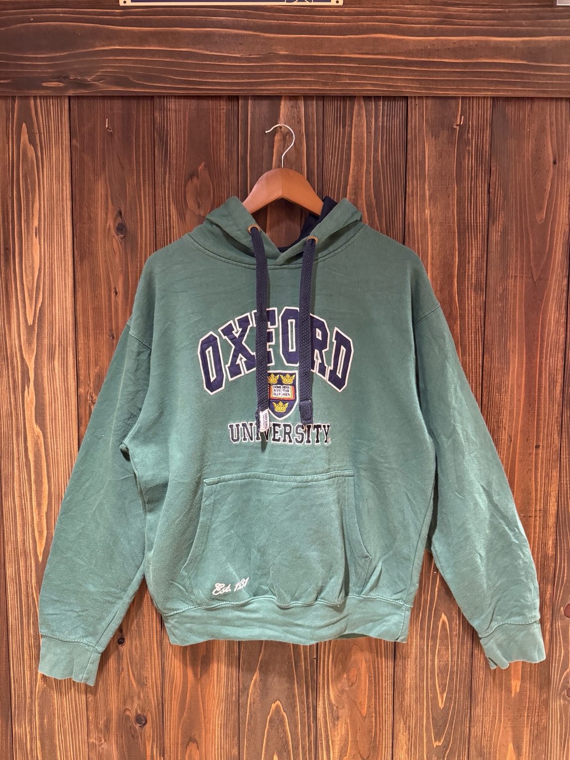 木目の壁に掛けたOXFORD UNIVERSITY公式グリーンパーカー（正面）