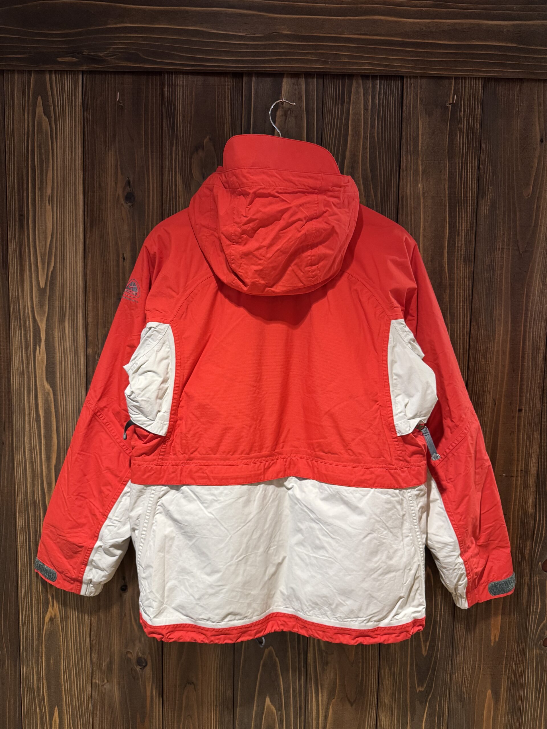 木の壁に掛けた赤×グレー切り替えのNIKE ACGシェルジャケット バックスタイル