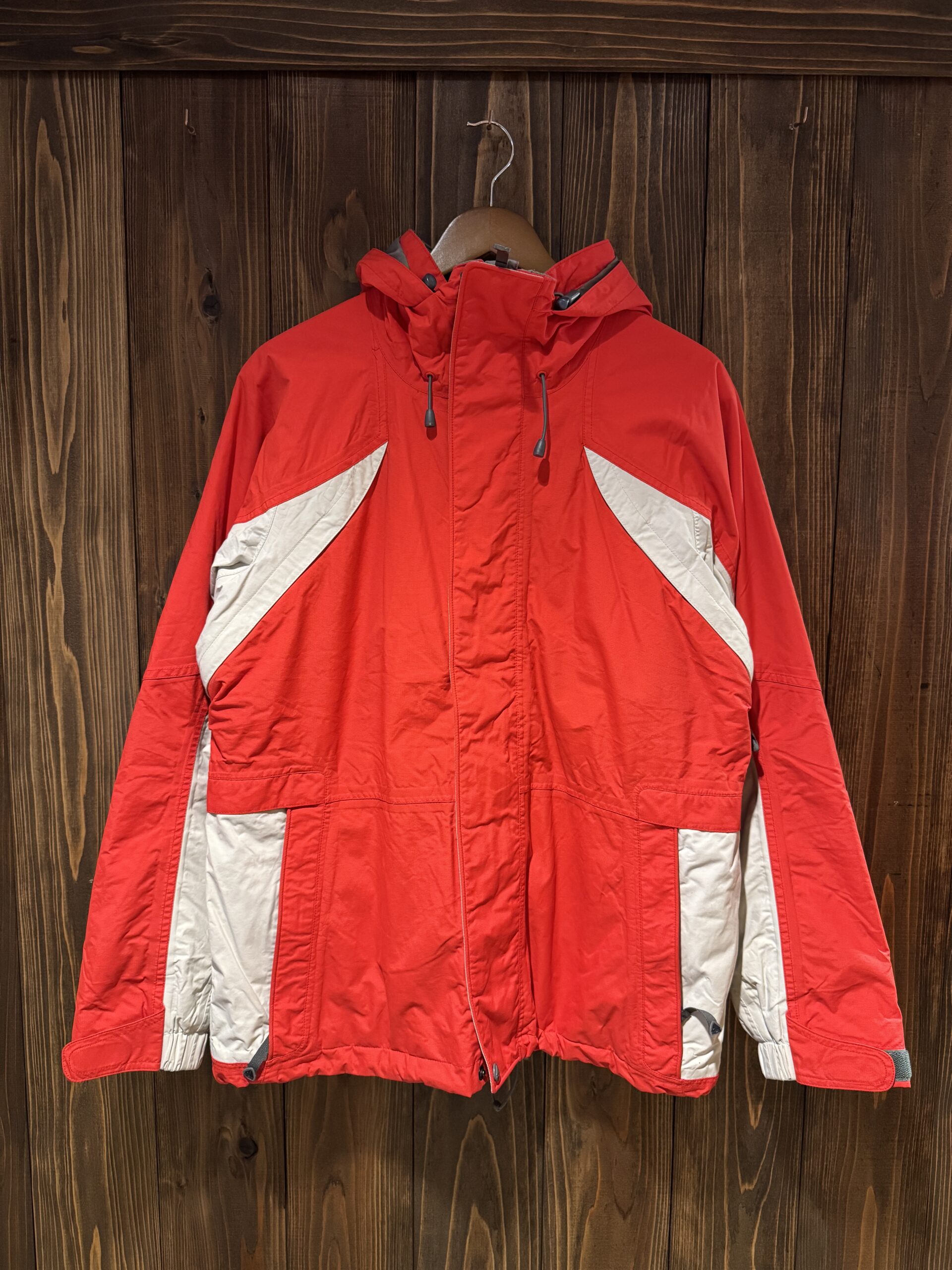 木の壁に掛けた赤×グレー切り替えのNIKE ACGシェルジャケット フロント全体