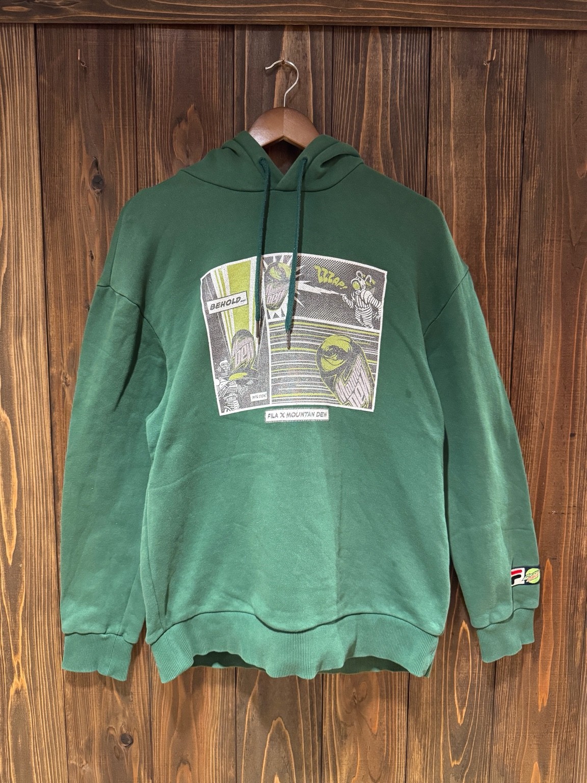 FILA × Mountain Dewのボトルグリーンフーディを店内の木製ハンガーに吊った正面全体写真。