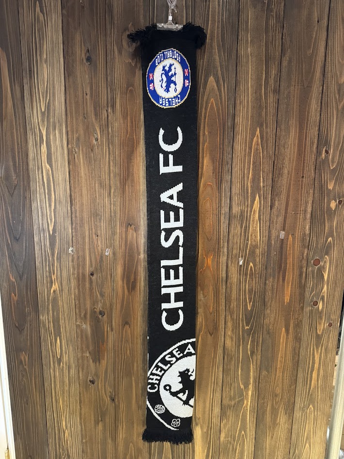 木の壁に吊るされたチェルシーFCのブラックサポーターマフラー。縦に大きくCHELSEA FCのロゴが編み込まれた全体画像。