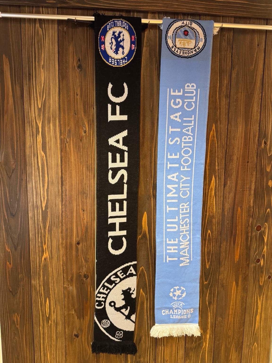 木の壁を背景に、チェルシーFCのブラック×ホワイトマフラーと、マンチェスター・シティのライトブルーマフラーを並べてハンガーパイプに掛けた様子。