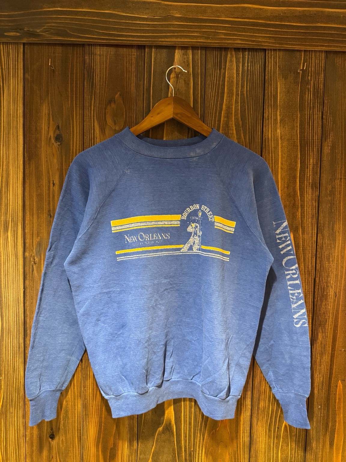 Healthknitのニューオリンズ土産スウェット。木壁背景でハンガー掛けした正面全体。Bourbon Street / New Orleansプリント。