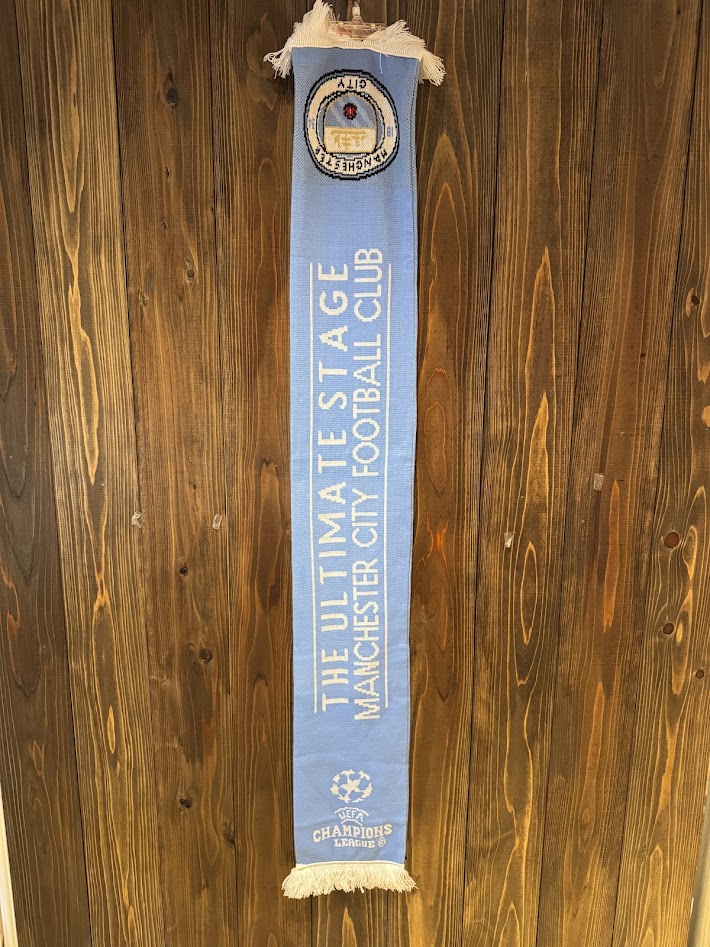 マンチェスター・シティ × UEFAチャンピオンズリーグ公式マフラーの全体（ハンガー掛け・フロント側）の様子。ライトブルーのボディにホワイトロゴが映える一枚。