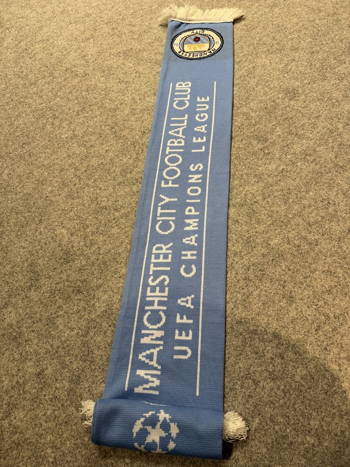 床に広げたマンチェスター・シティ × UEFAチャンピオンズリーグ公式マフラー。MANCHESTER CITY FOOTBALL CLUB と UEFA CHAMPIONS LEAGUE のロゴが並ぶデザイン。
