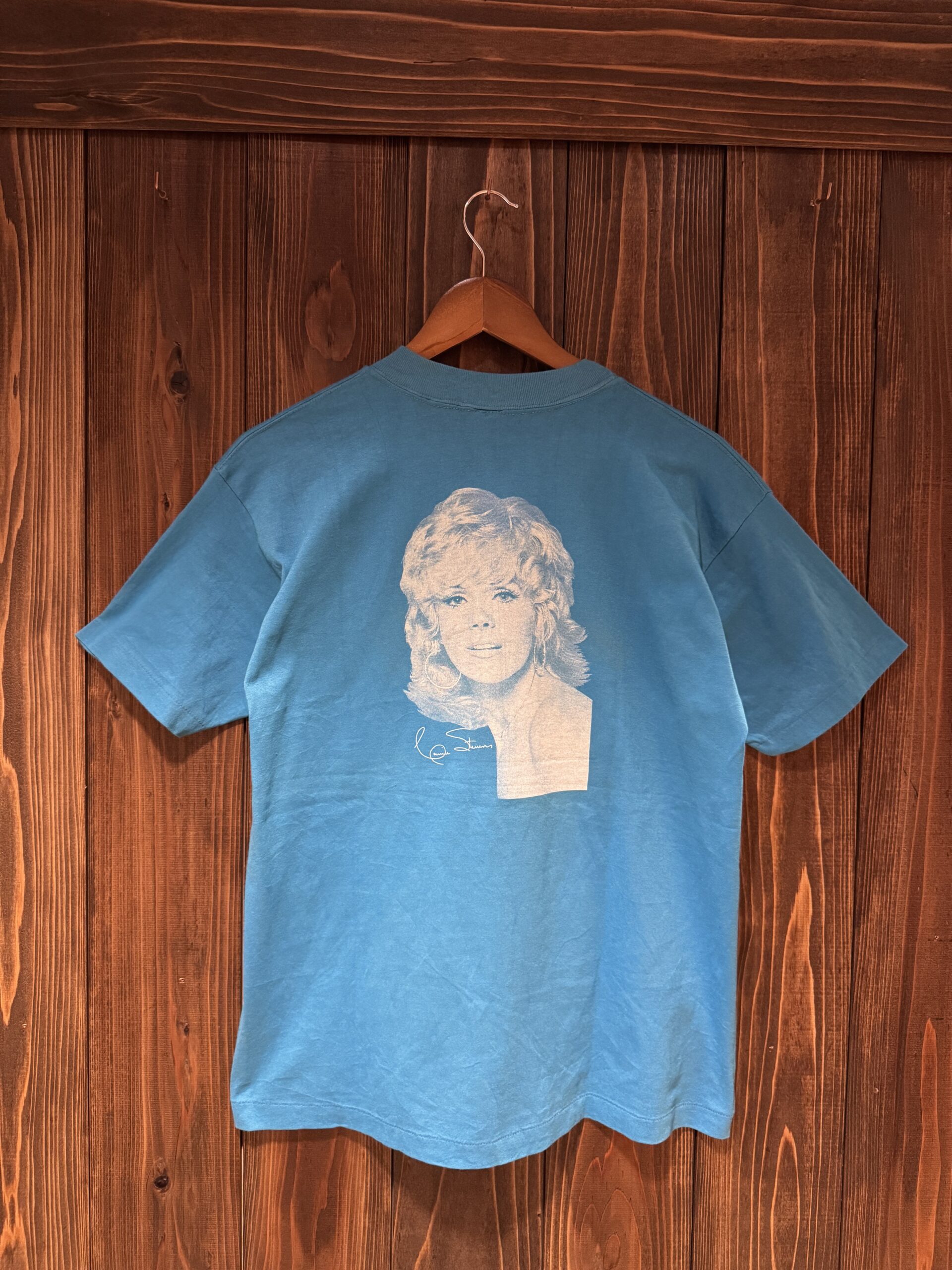 バックに女性のポートレートプリントが入ったWINDFEATHER SUMMER CAMPのTシャツ。木の壁にハンガー掛けでディスプレイされています。